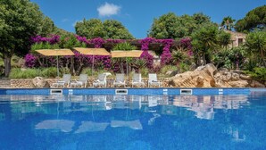 Une piscine d’un bleu éclatant, bordée de chaises longues et d’ombrelles, et entourée d’une végétation luxuriante et de fleurs violettes.