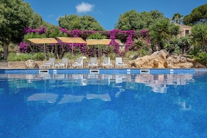 Una piscina blava cristal·lina amb gandules i ombrel·les, envoltada de vegetació exuberanta i flors morades.