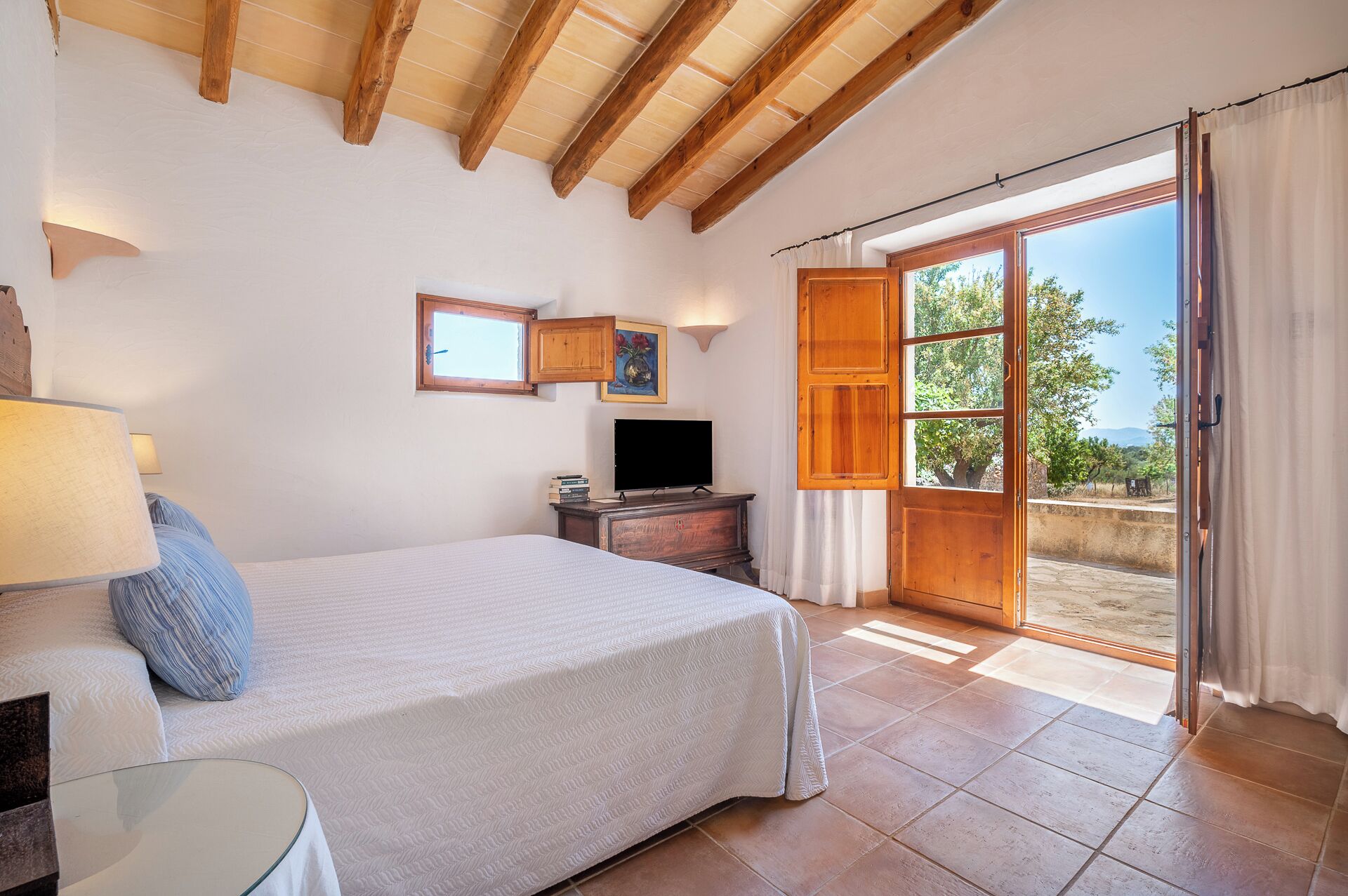 A bedroom with a bed, a TV, a small table, a lamp, a window, and a door leading to an outdoor area.