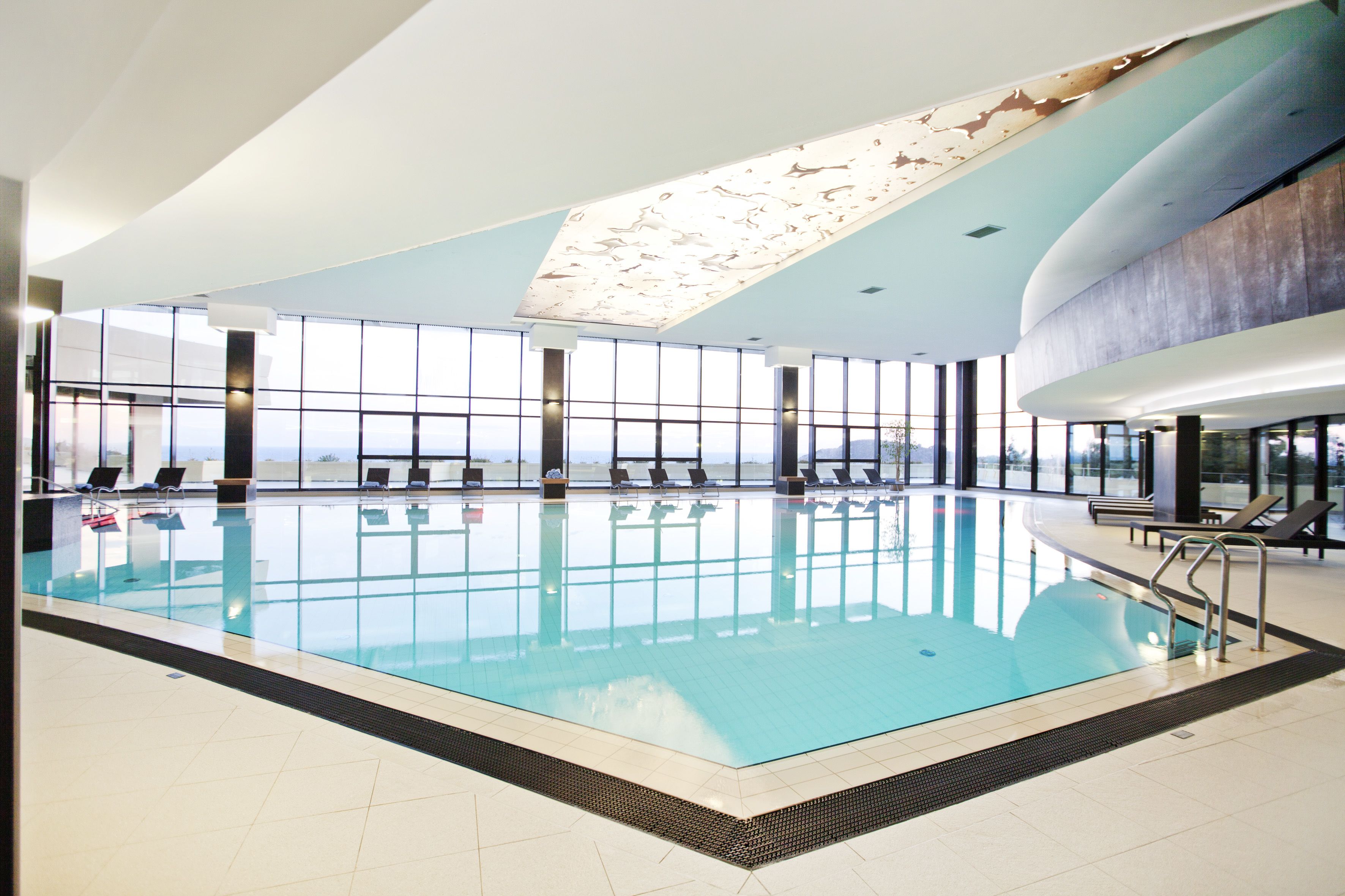 Une grande piscine intérieure à l’eau d’un bleu cristallin, entourée de chaises longues et dotée d’un plafond haut orné d’un luminaire original.