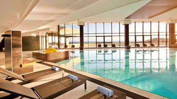 An indoor swimming pool with lounge chairs and a view of the sea.