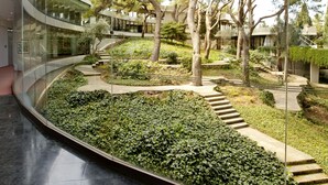 Ein modernes Gebäude mit einer geschwungenen Glasfassade, einem begrünten Garten mit Bäumen und einem Weg sowie einer Treppe, die zu einer oberen Ebene führt.