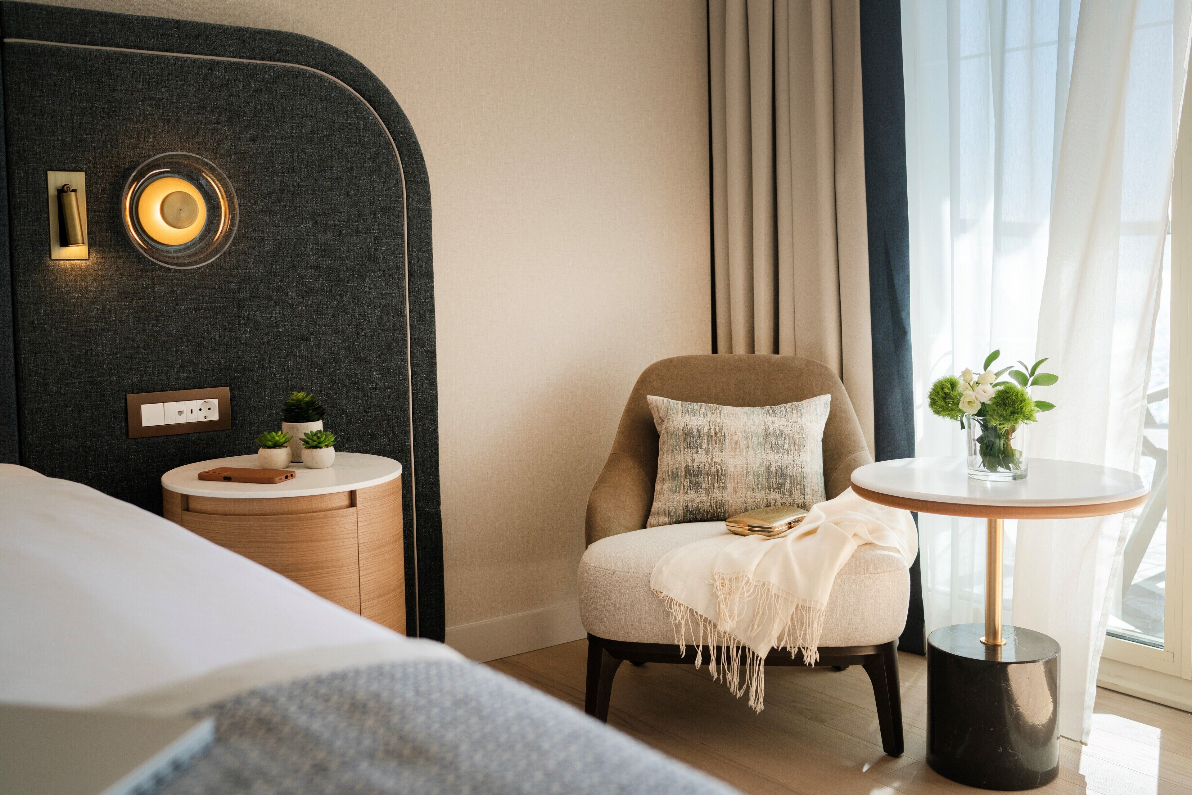 A modern hotel room with a bed, a round table with a vase of flowers, a chair with a cushion, a small side table with a phone and plants, and a wall-mounted light fixture.