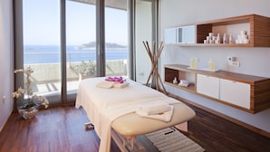 Ein moderner Spa-Raum mit einer Massageliege, Holzregalen mit Pflegeprodukten und einem großen Fenster mit Meerblick.