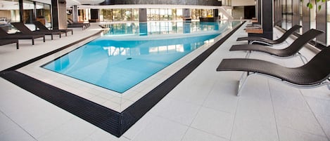 An indoor swimming pool with lounge chairs, a tiled floor, and large windows.