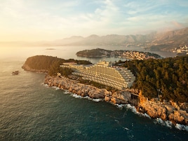 Un hotel costero con varias construcciones, un litoral rocoso y vistas al mar y montañas distantes.