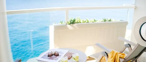 Un balcón con vista al mar, una mesa con bebidas y gafas de sol, y una silla con una toalla amarilla.