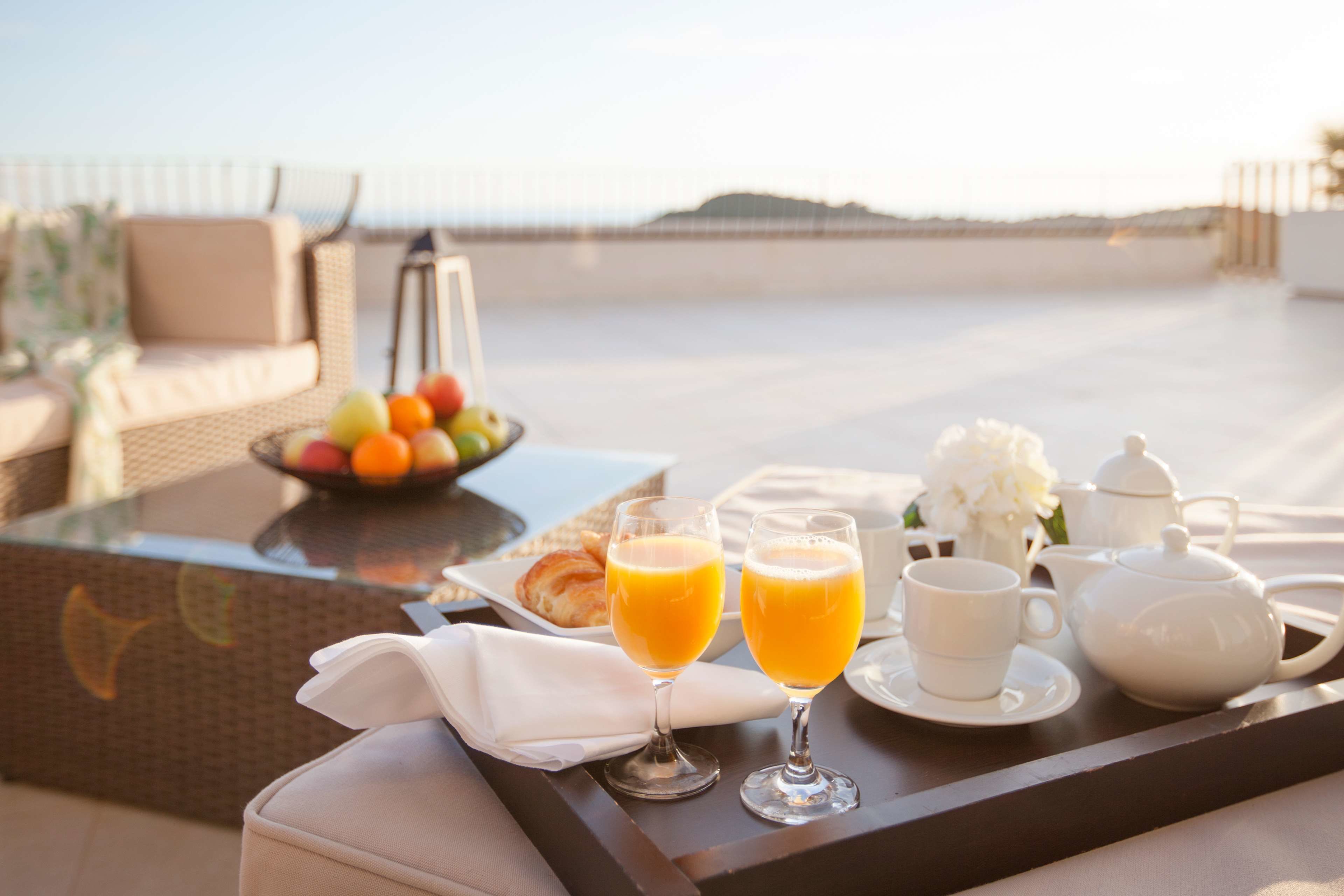 A breakfast tray with orange juice, a teapot, and a cup on a table with a wicker chair and a bowl of fruit in the background.