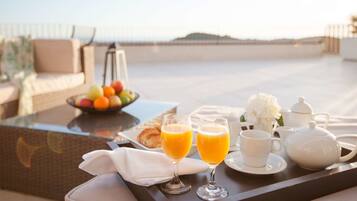 Bandeja de café da manhã com suco de laranja, um bule e uma xícara sobre uma mesa, com uma cadeira de vime e uma tigela de frutas ao fundo.