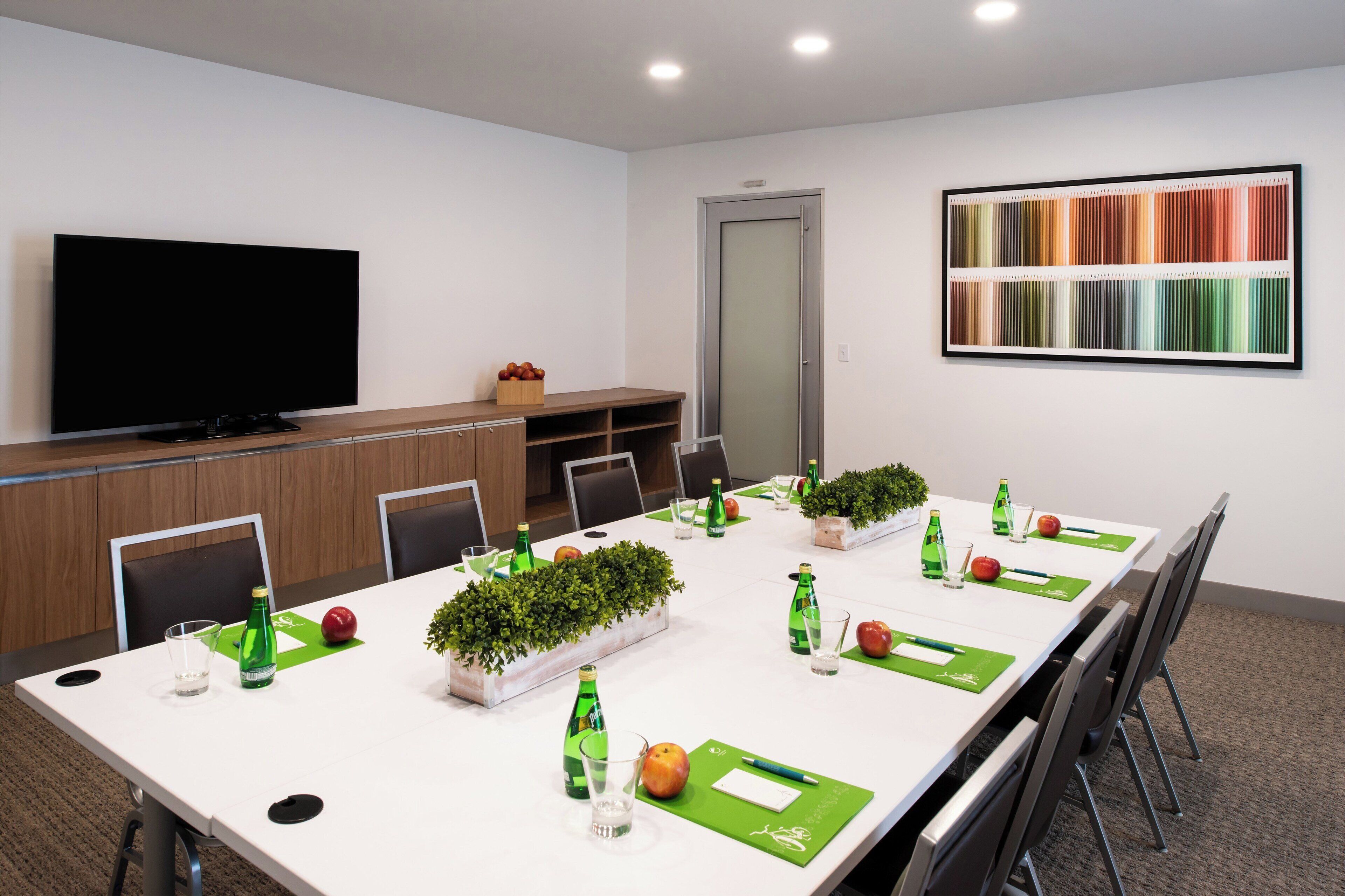 Uma sala de conferências com uma mesa comprida, cadeiras, uma televisão e uma paleta de cores na parede.