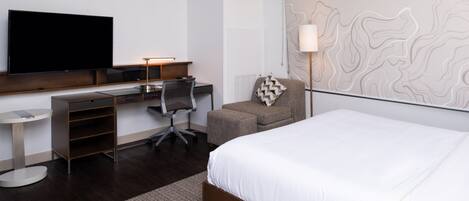 A modern hotel room with a large bed, a desk with a computer, a flat-screen TV, and a wall-mounted lamp.