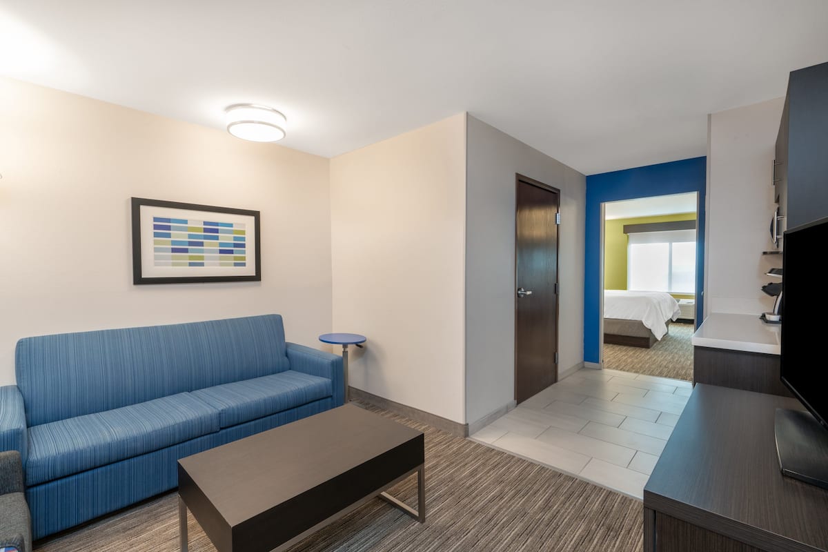 A hotel room with a blue sofa, a small table, a framed picture on the wall, a door leading to a bathroom, and a TV on a stand.