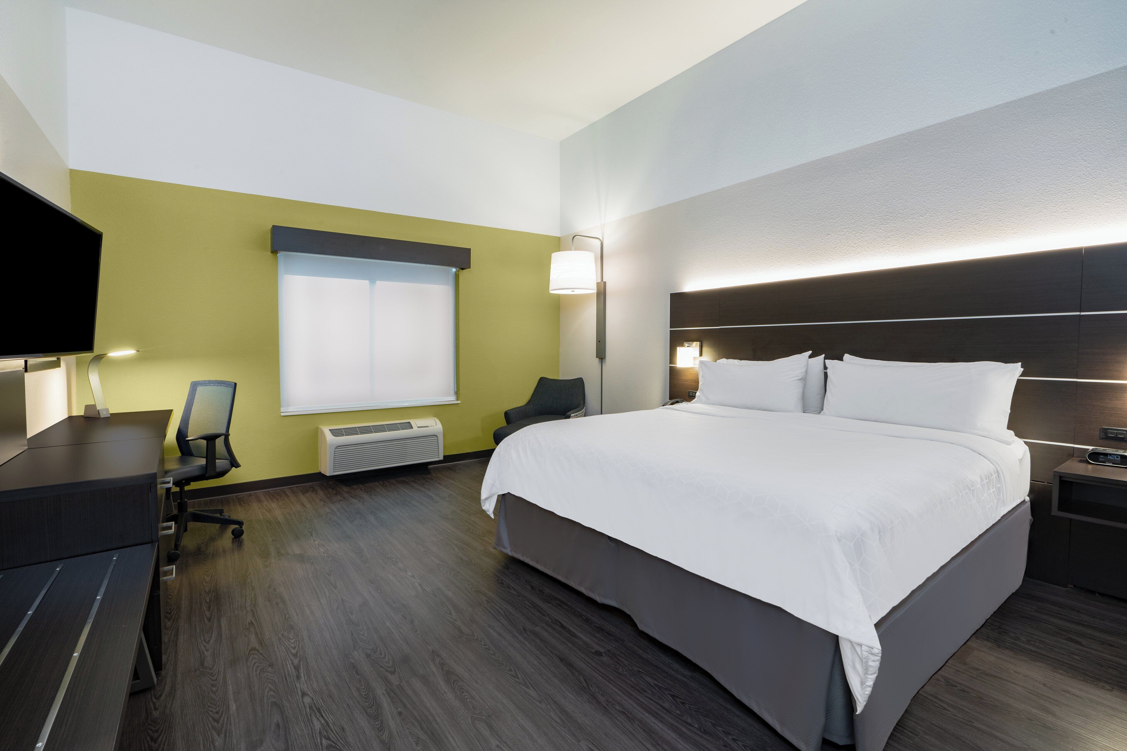A hotel room with a large bed, a desk, a chair, a TV, and a window with blinds.