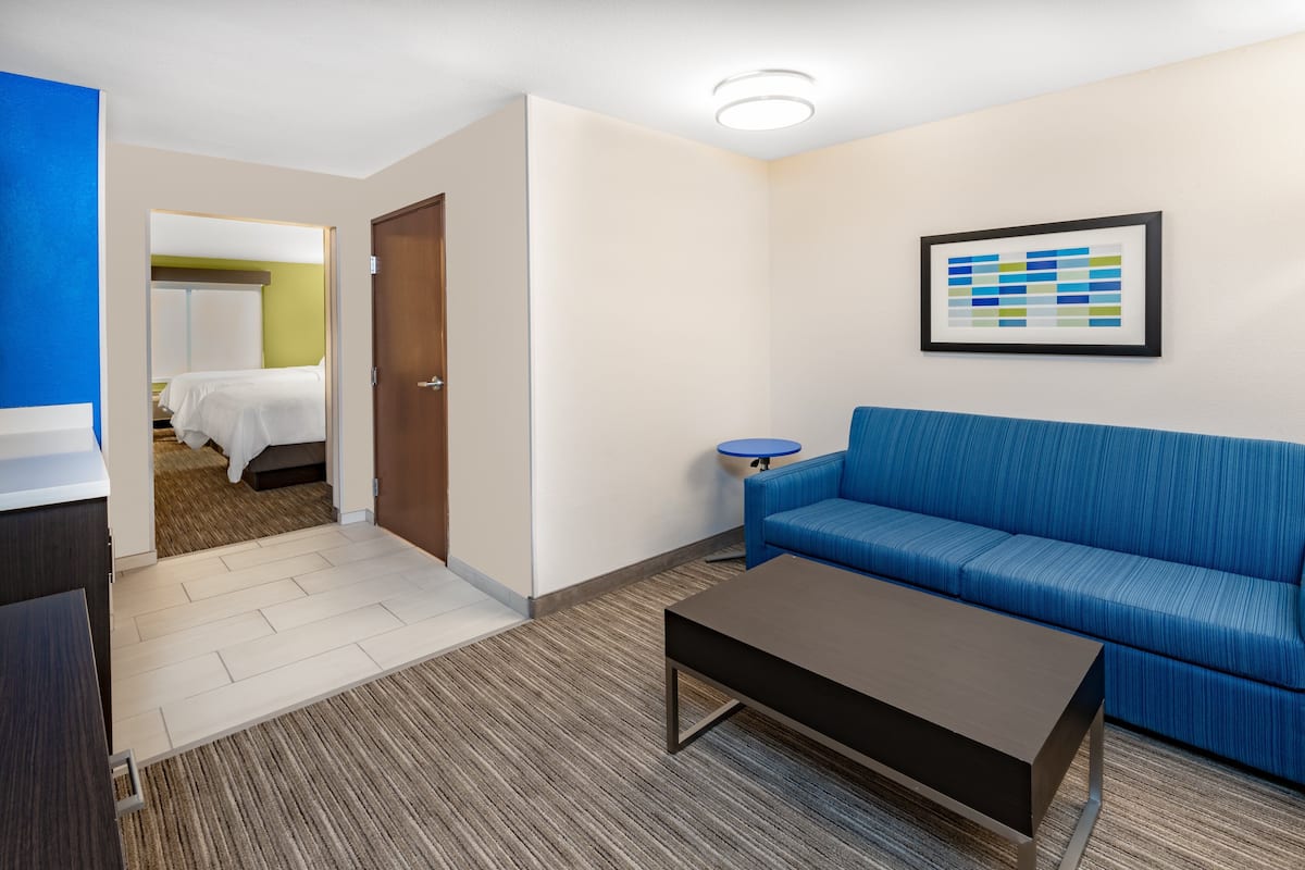A modern hotel room with a blue sofa, a coffee table, a framed artwork on the wall, and a door leading to a bedroom.