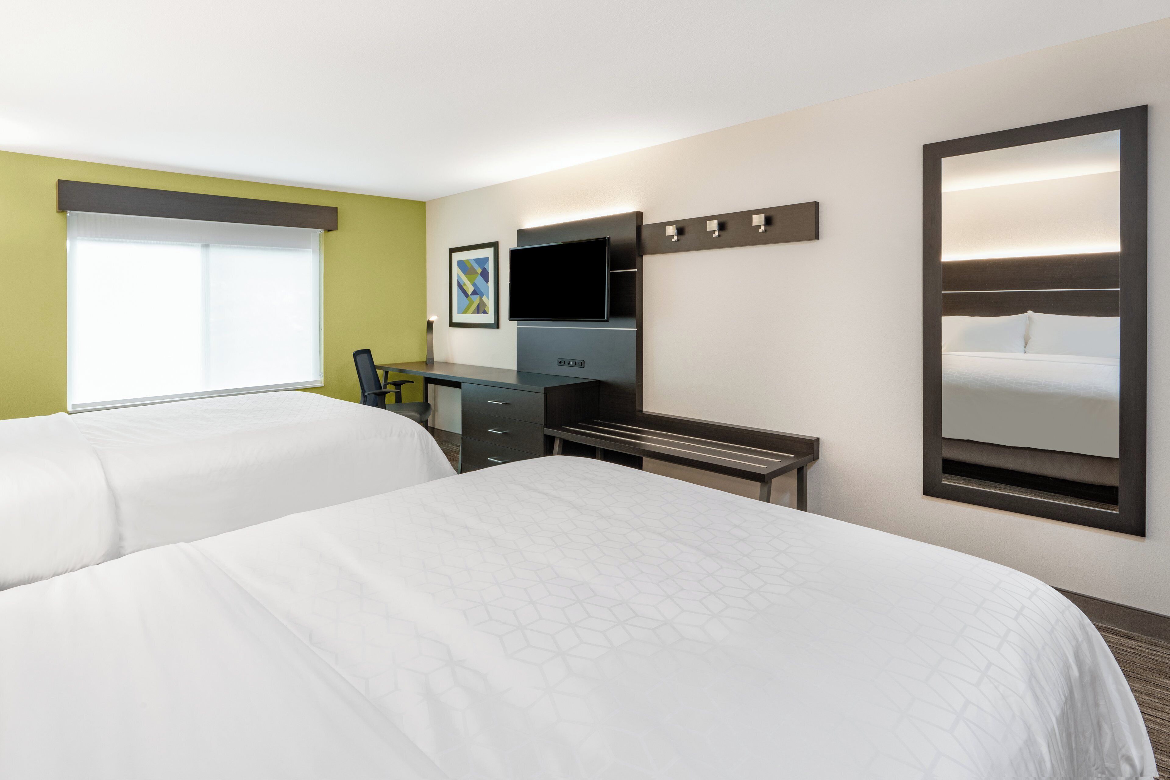 Habitación de hotel con dos camas, un escritorio, un televisor de pantalla plana, una silla y un espejo grande.
