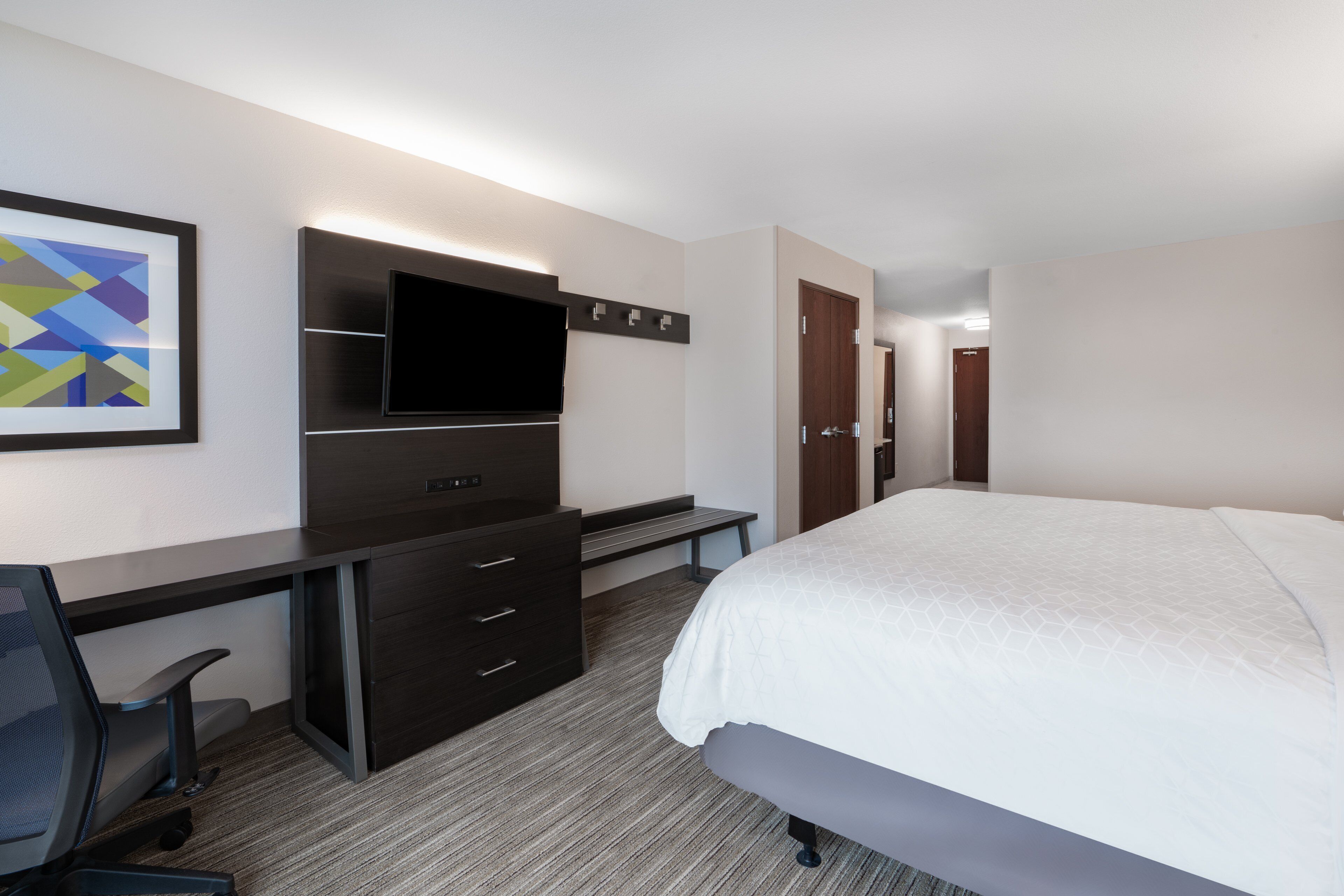 Une chambre d’hôtel comprenant un lit, un bureau avec une chaise, un téléviseur à écran plat, une commode et un tableau encadré au mur.