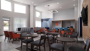 Een moderne hotel lobby met meerdere zithoeken, waaronder tafels en stoelen, en een groot televisiescherm aan de muur.