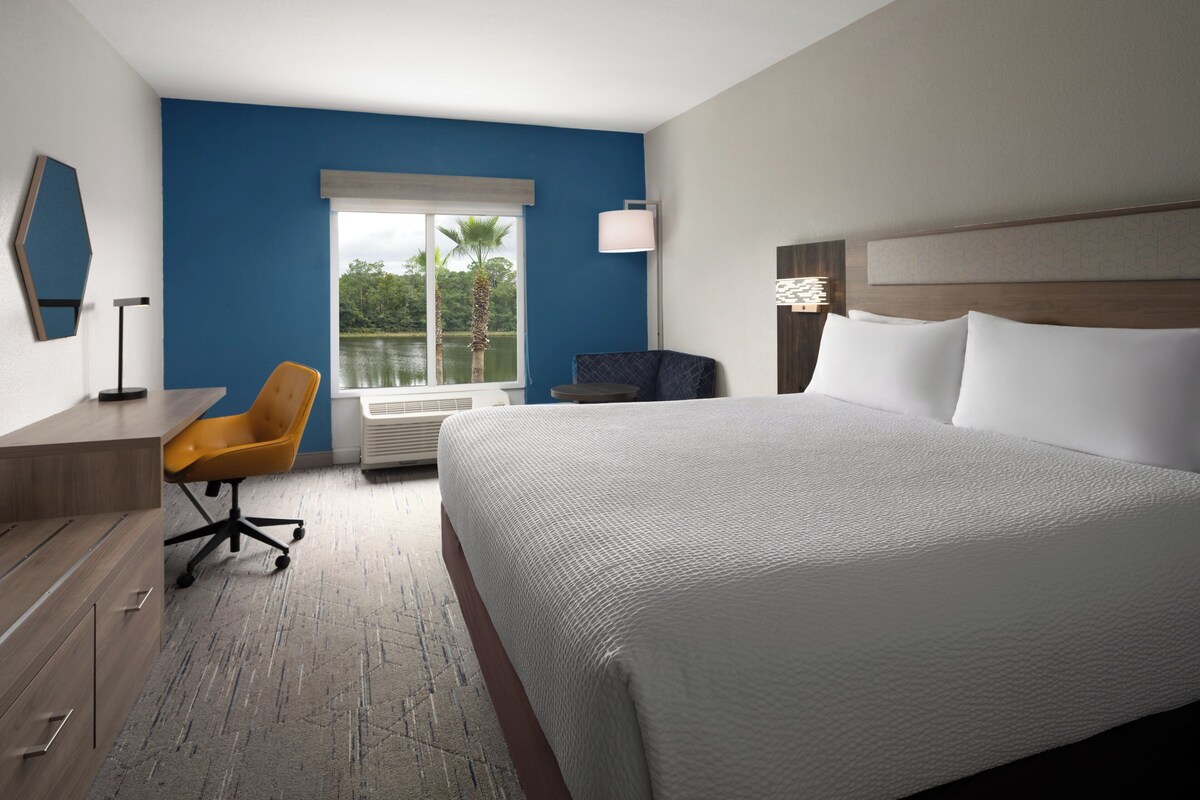 Een hotelkamer met een groot bed, een bureau met stoel, een raam met uitzicht op bomen en een lamp.