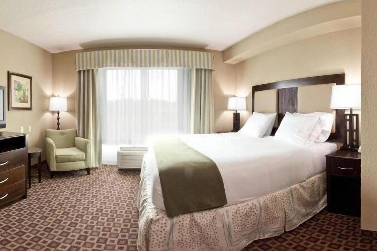 Une chambre d’hôtel avec un grand lit, deux tables de chevet avec des lampes, une chaise, un bureau et une grande fenêtre avec des rideaux.