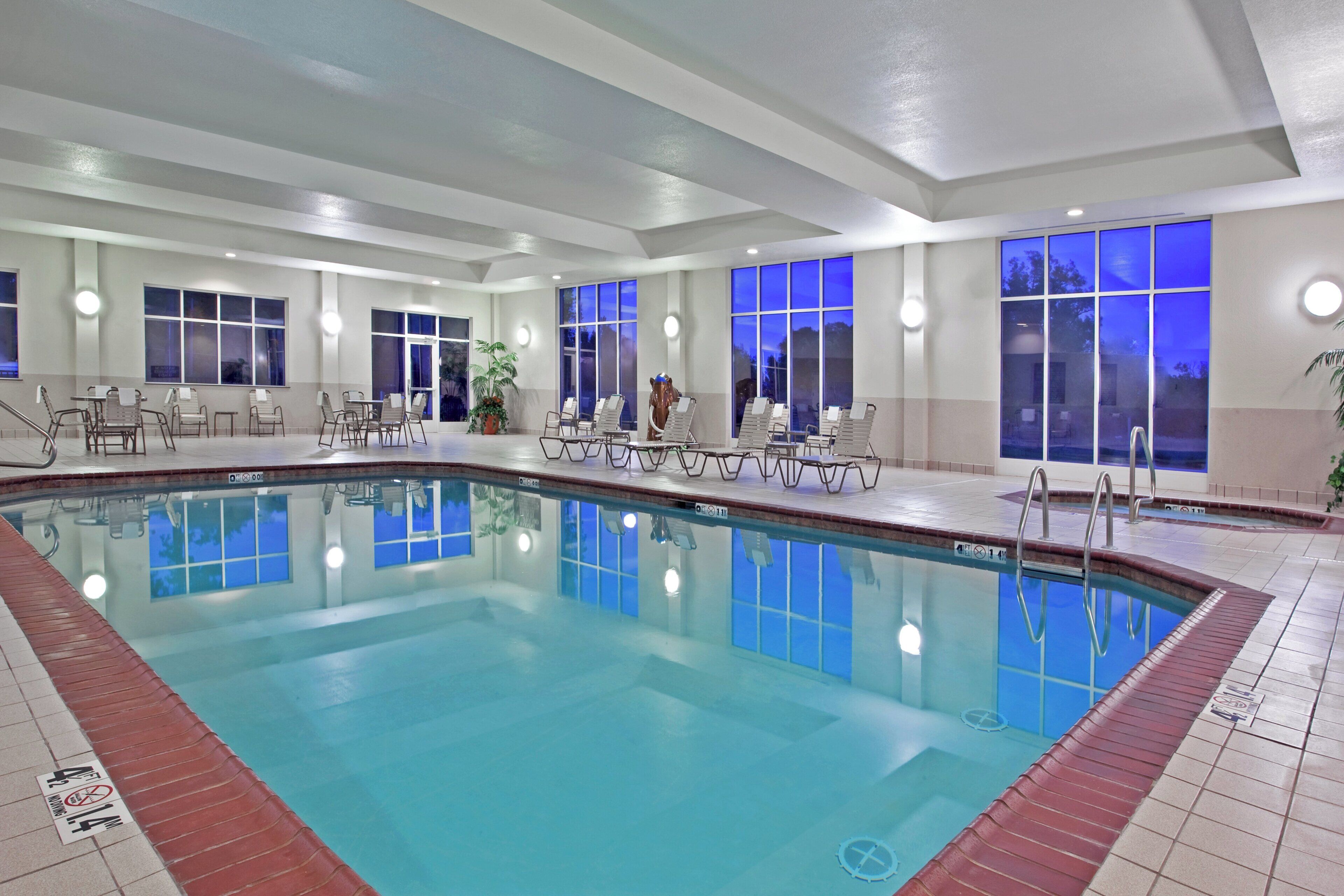 A hotel indoor pool with clear water, surrounded by lounge chairs and tables.