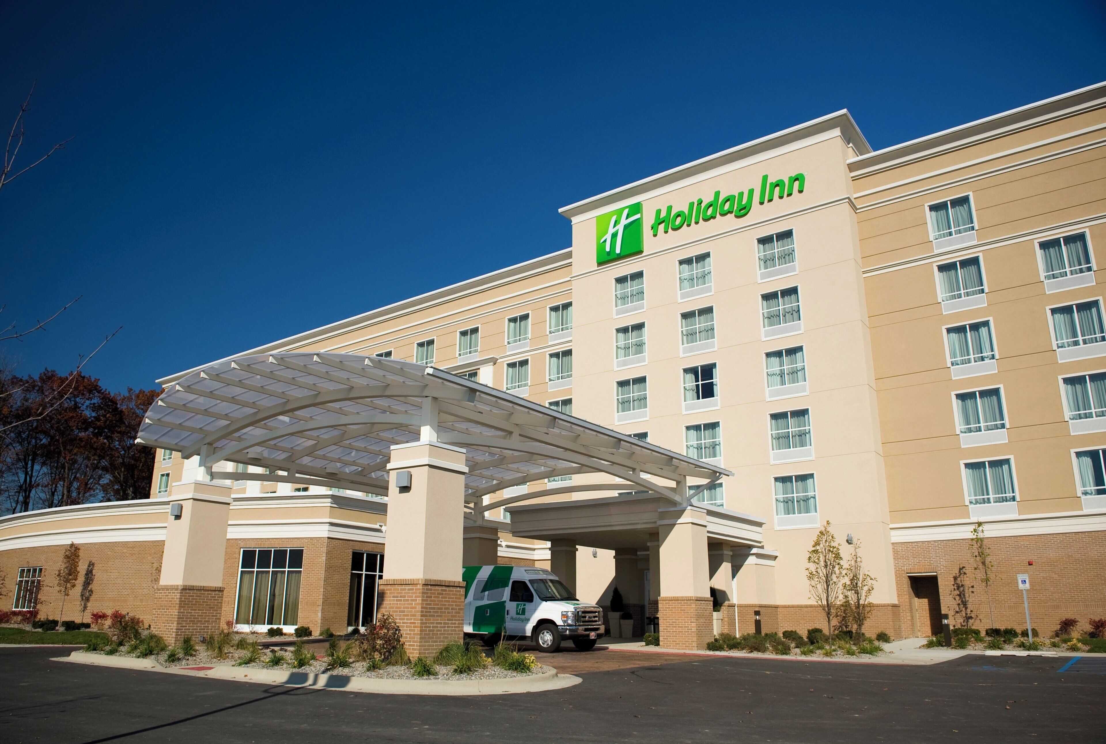 Um hotel Holiday Inn com entrada coberta, estacionamento e um ônibus de transporte verde e branco.
