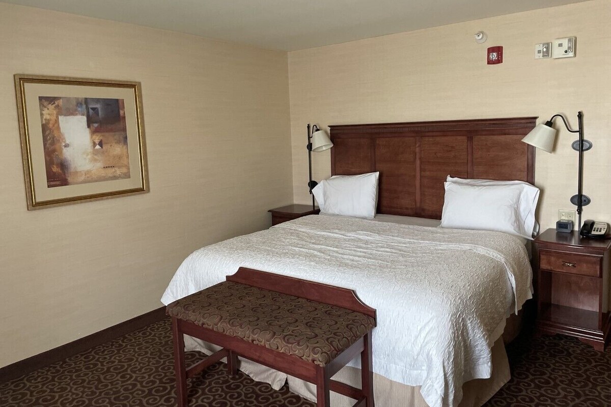A hotel room with a large bed, a wooden headboard, a bedside table with a lamp, a bench, and a framed picture on the wall.