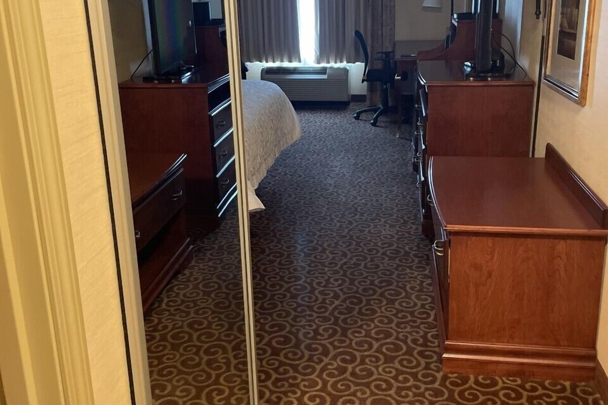A hotel room with a bed, a desk, a chair, a TV, a lamp, and a mirror.