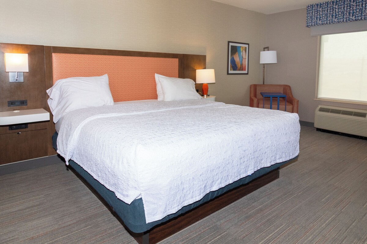 A hotel room with a large bed, a nightstand, a chair, and a window.
