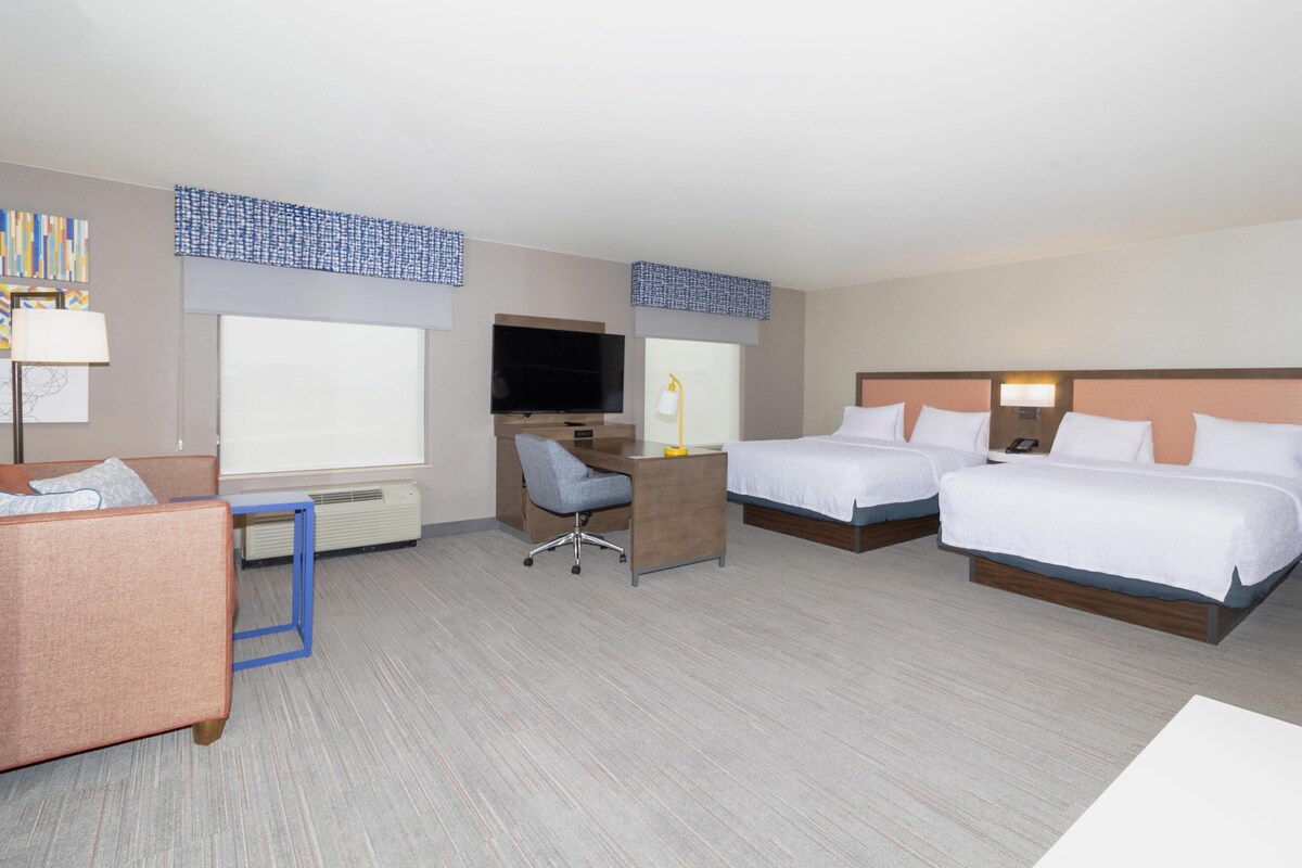 A hotel room with a desk, two beds, a TV, and a window.