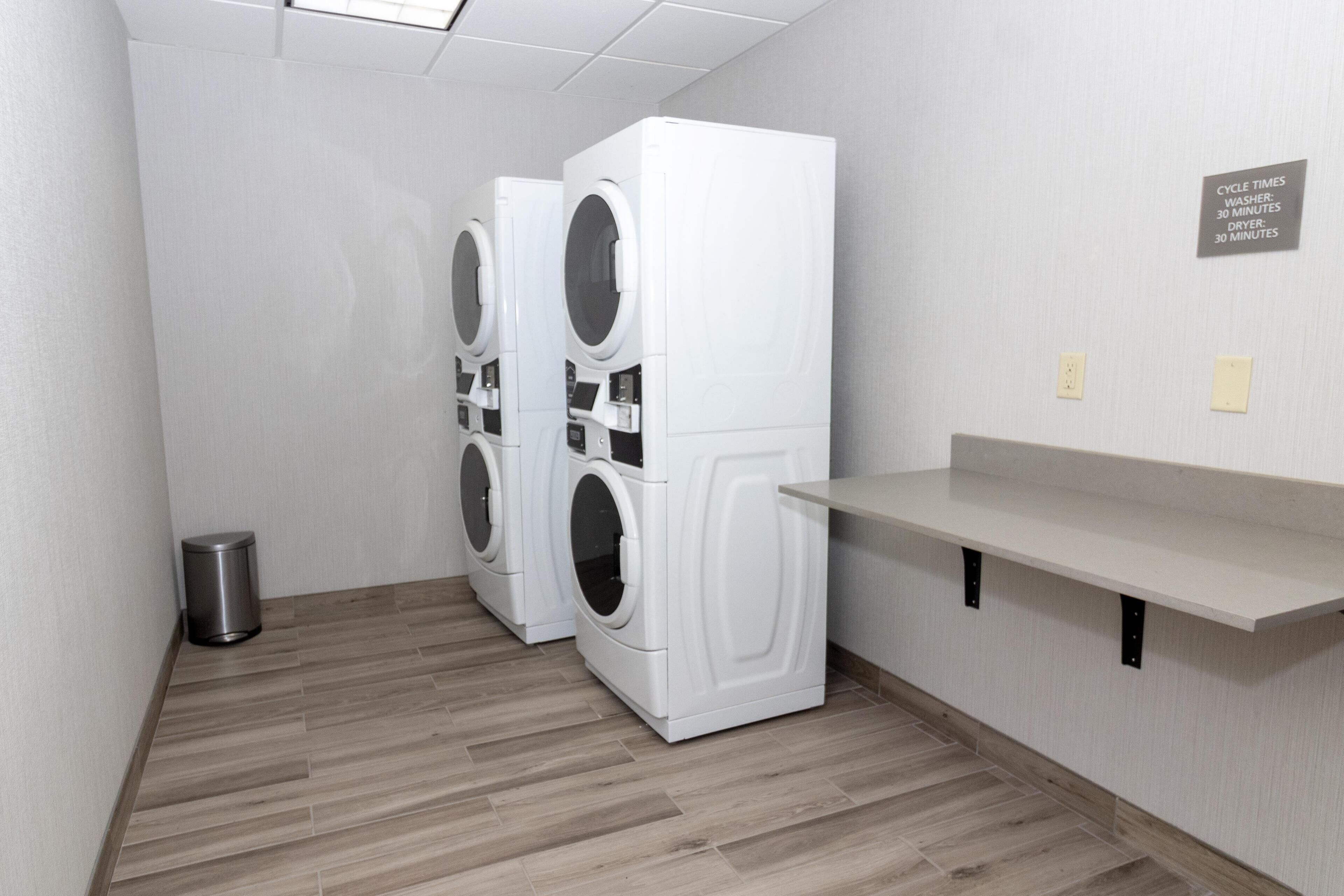 A laundry room with three stacked washers and dryers, a metal trash can, and a wall-mounted shelf.
