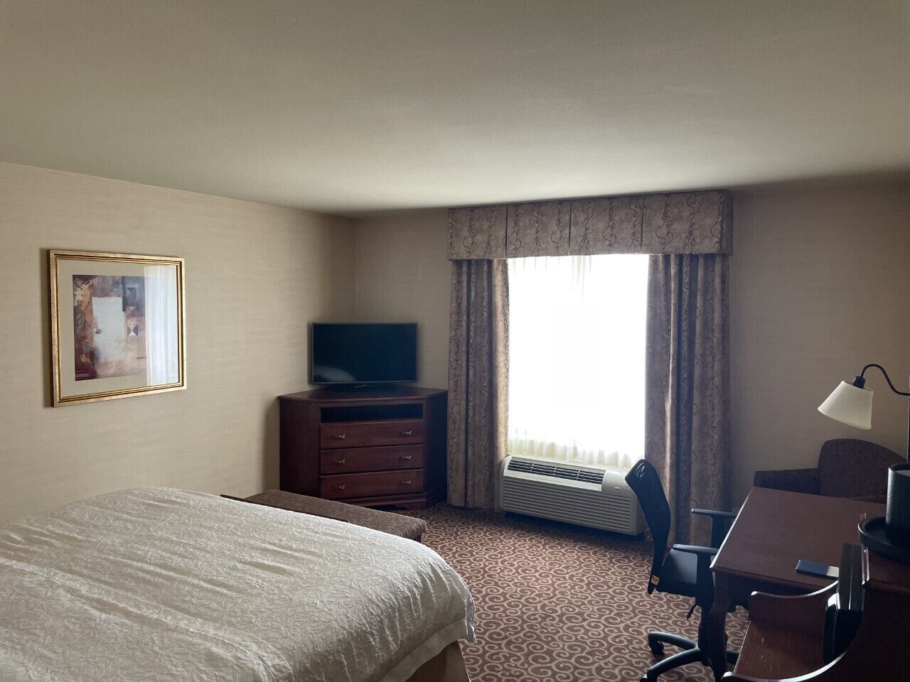 A hotel room with a bed, a desk with a chair, a television, a dresser, and a window with curtains.