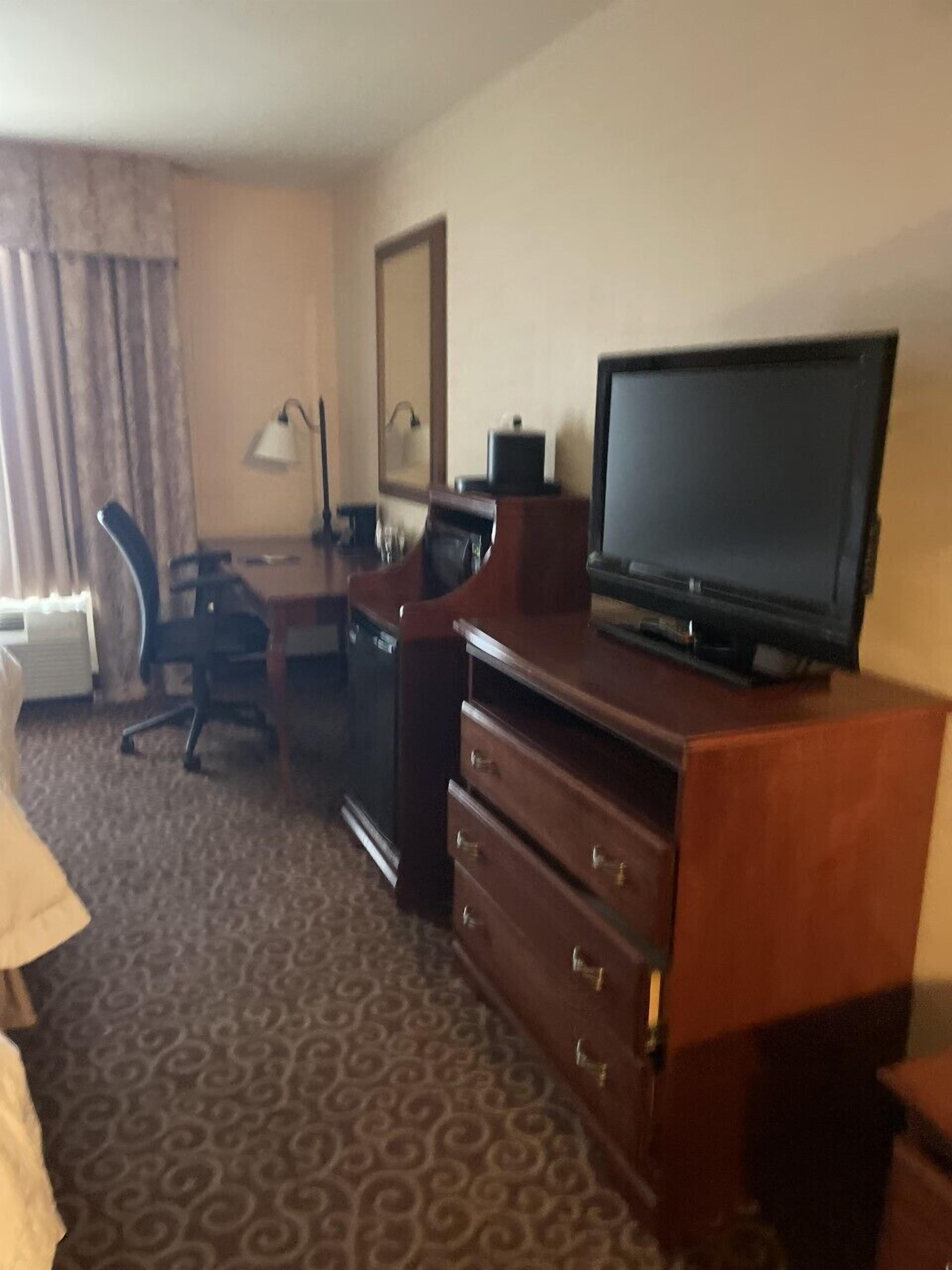 A hotel room with a television on a wooden stand, a desk with a chair, a dresser, and a patterned carpet.
