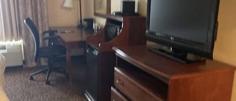 A hotel room with a television on a wooden stand, a desk with a chair, a dresser, and a patterned carpet.