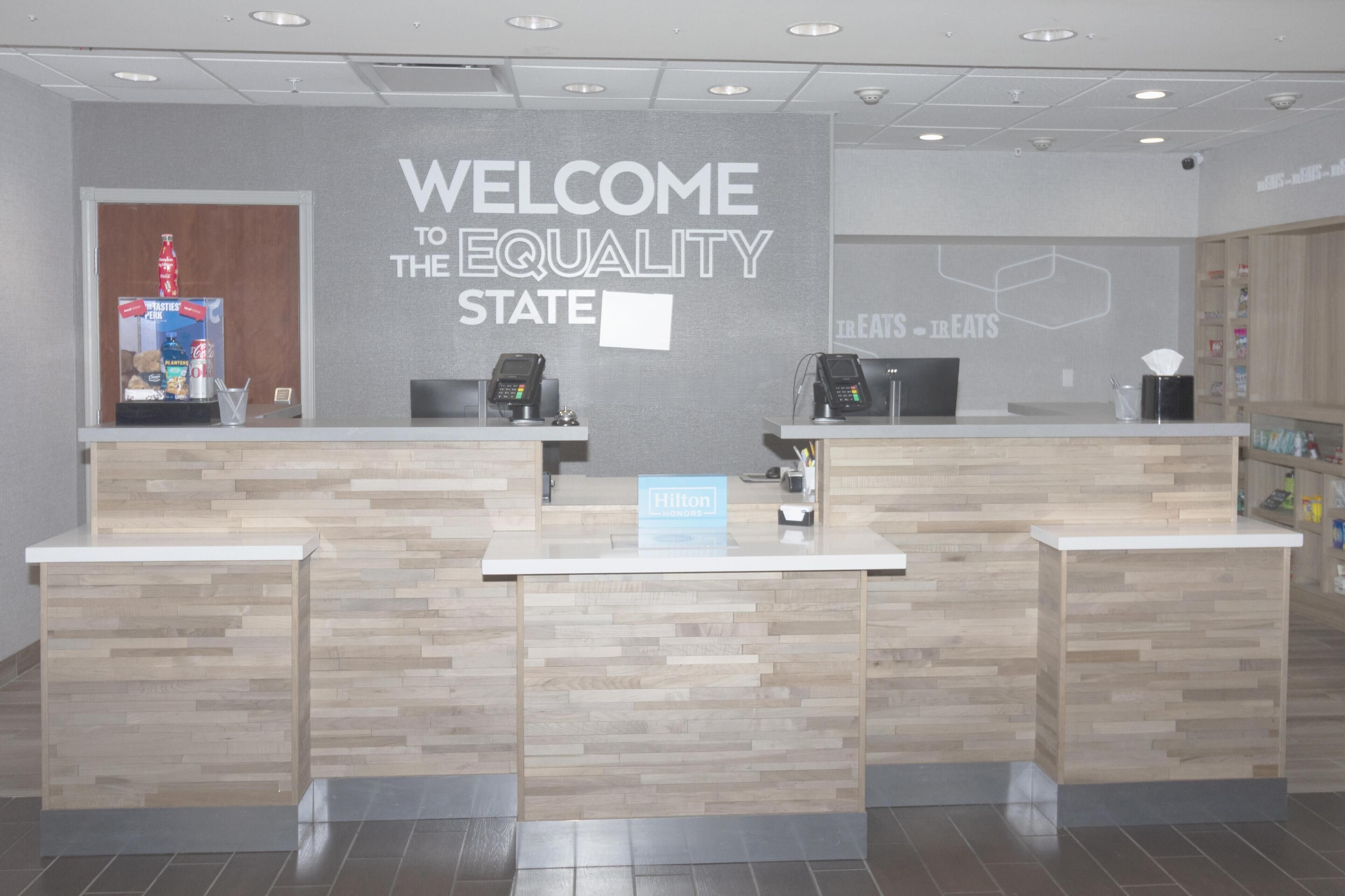 A reception desk with a 'WELCOME TO THE EQUALITY STATE' sign, two card readers, and a 'HOTEL' sign.
