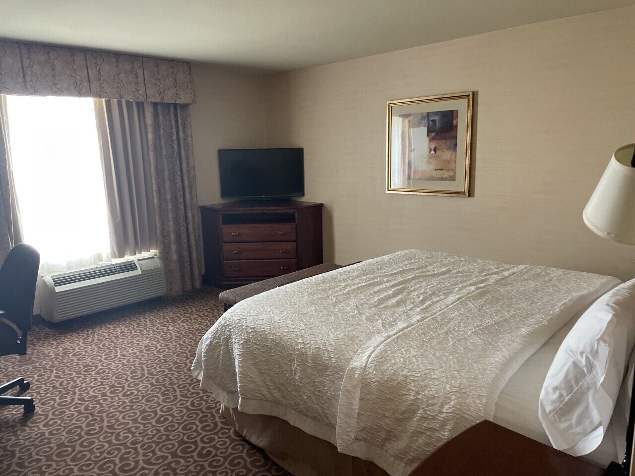 A hotel room with a bed, a television on a wooden dresser, a framed picture on the wall, and a window with curtains.