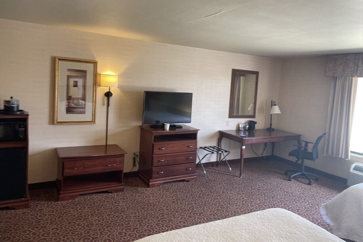 A hotel room with a bed, a desk with a chair, a television on a dresser, and a lamp.