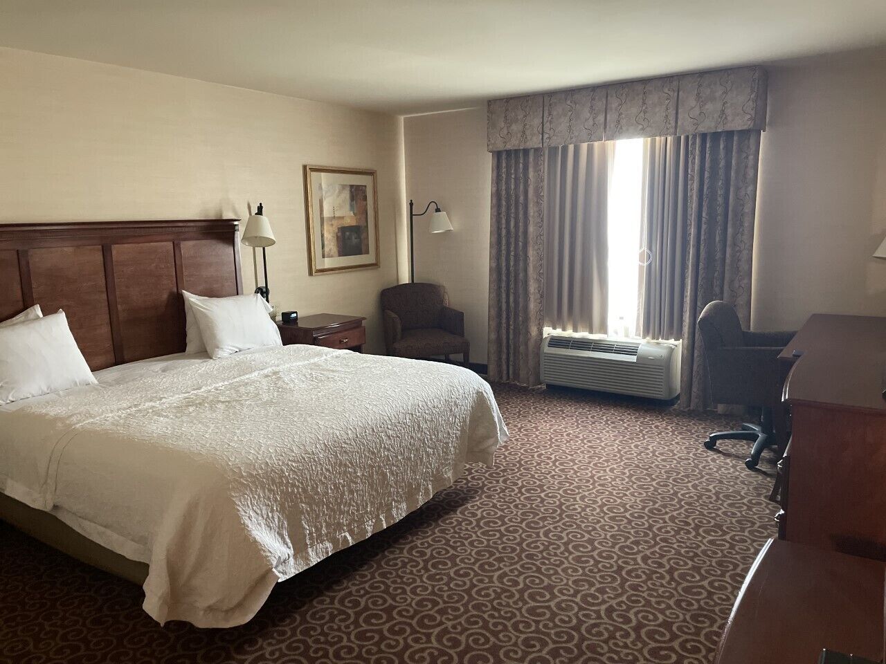 A hotel room with a large bed, a desk, a chair, a lamp, a picture on the wall, and a window with curtains.