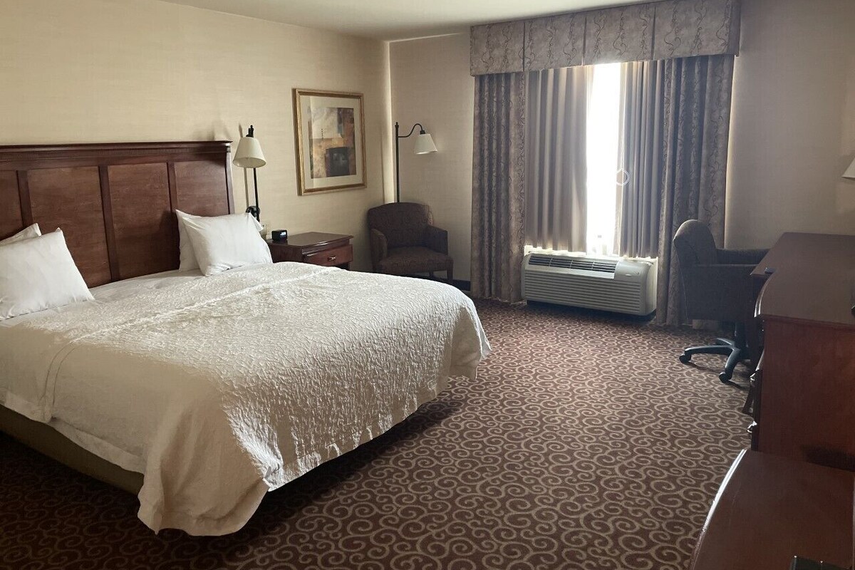 A hotel room with a large bed, a desk, a chair, a lamp, a picture on the wall, and a window with curtains.