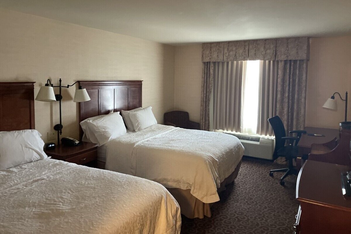 A hotel room with two beds, a desk, a chair, a lamp, and a window with curtains.