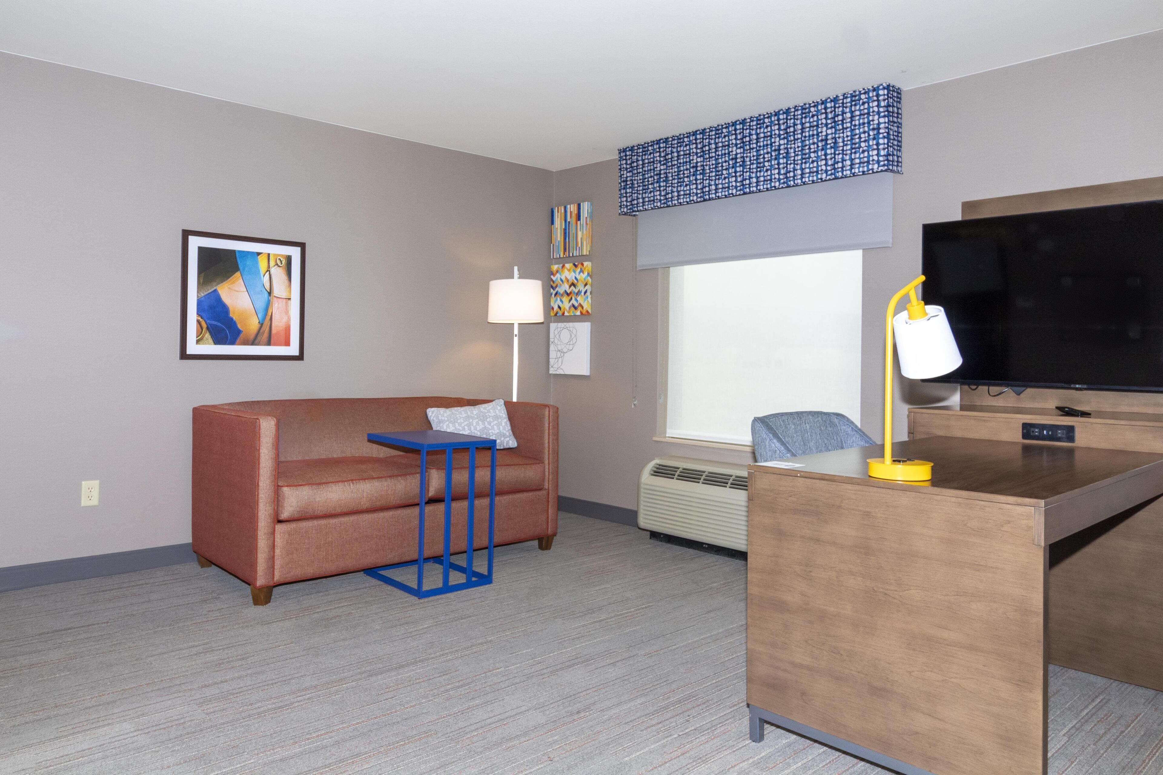 A hotel room with a sofa, a small table, a desk with a lamp, a TV, and artwork on the walls.