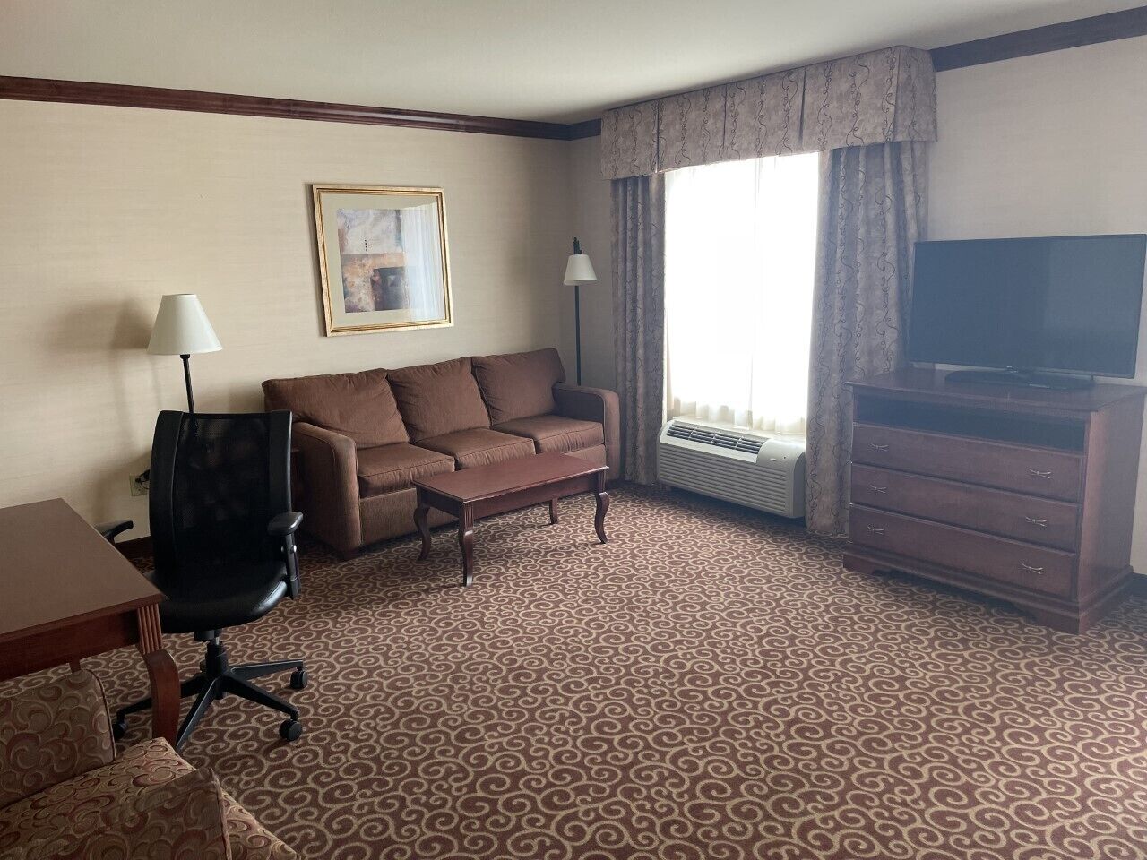 A hotel room with a brown patterned carpet, a brown sofa, a black office chair, a wooden dresser, a flat-screen TV, and a framed picture on the wall.
