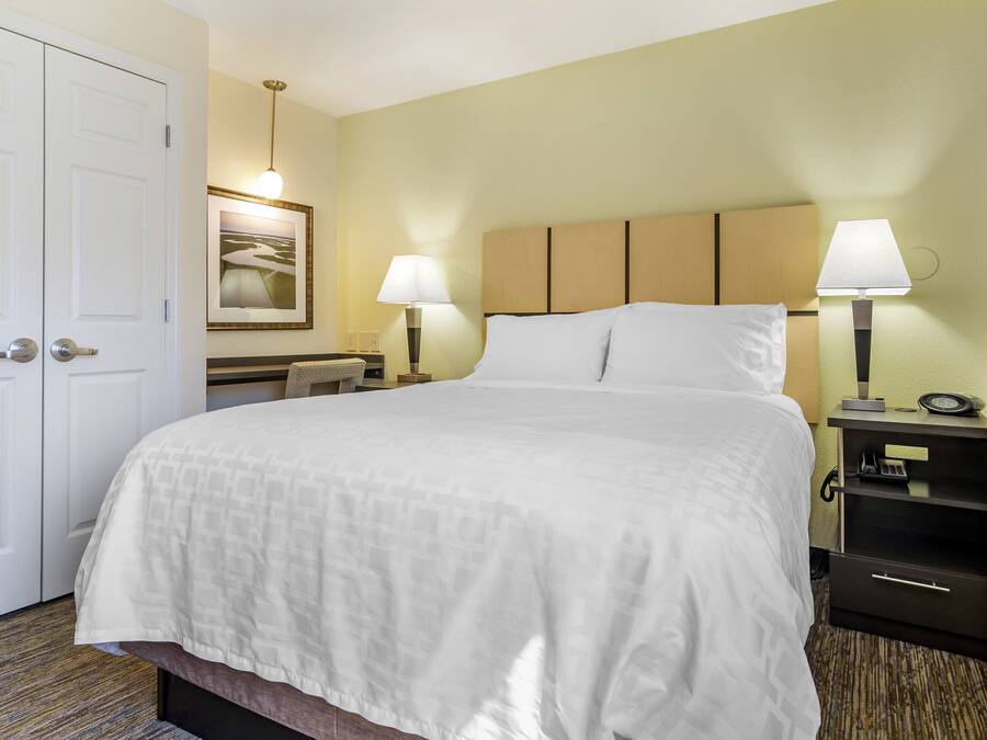 A hotel room with a large bed, two bedside lamps, a desk with a chair, a framed picture on the wall, and a door.