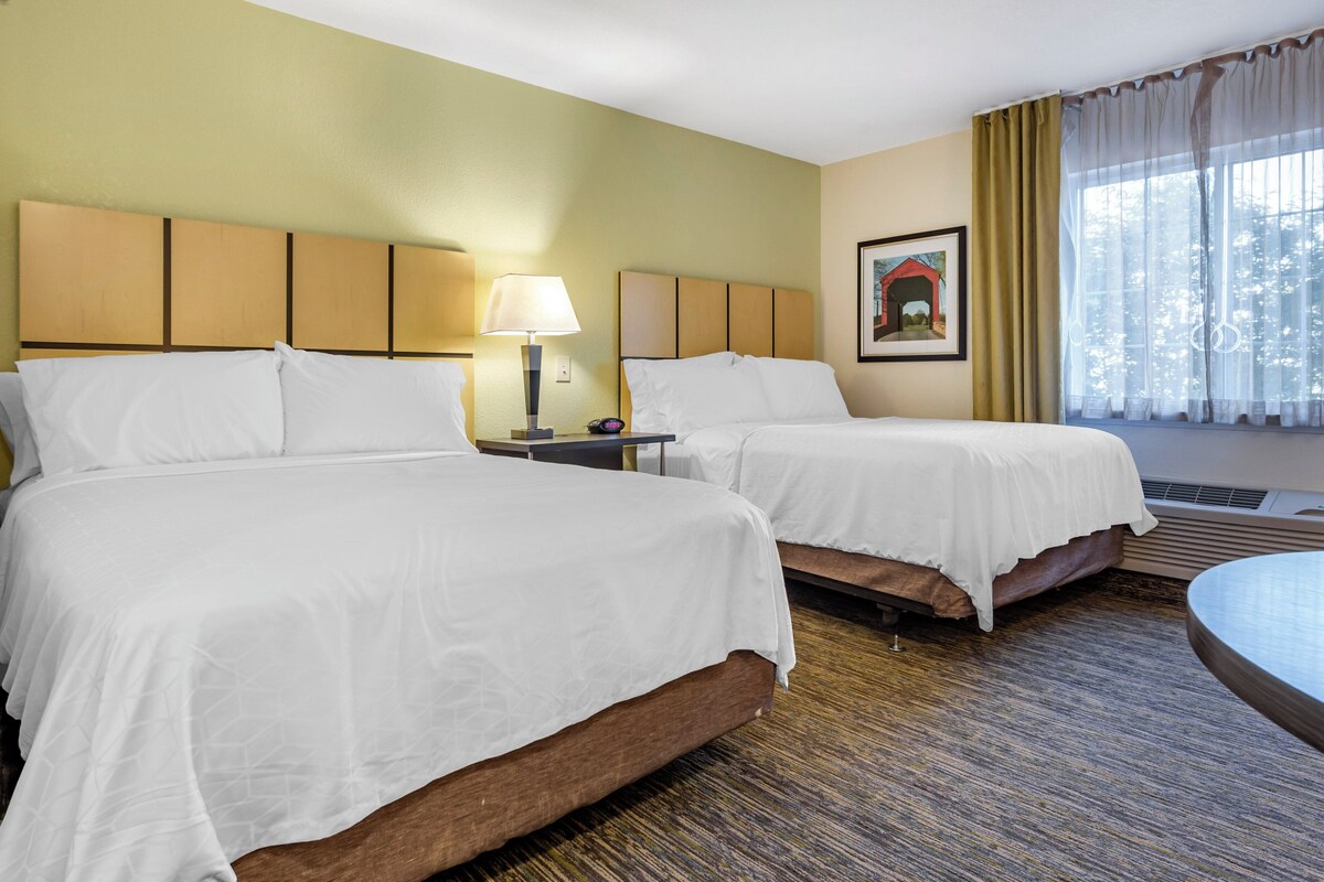 Een hotelkamer met twee bedden, een nachtkastje met een lamp, een lijst met een afbeelding aan de muur, en een raam met gordijnen.