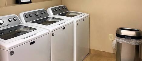 A laundry area with three washers and dryers, a trash can, and a framed picture on the wall.