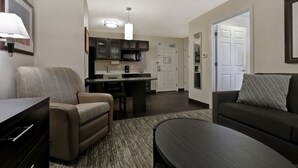 A modern living room with a sofa, a coffee table, and a dining area with a kitchen in the background. - Candlewood Suites Indianapolis - South by IHG (Greenwood)