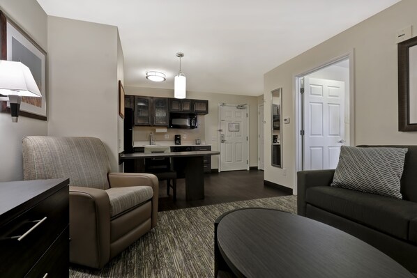 A modern living room with a sofa, a coffee table, and a dining area with a kitchen in the background. - Candlewood Suites Indianapolis - South by IHG (Greenwood)