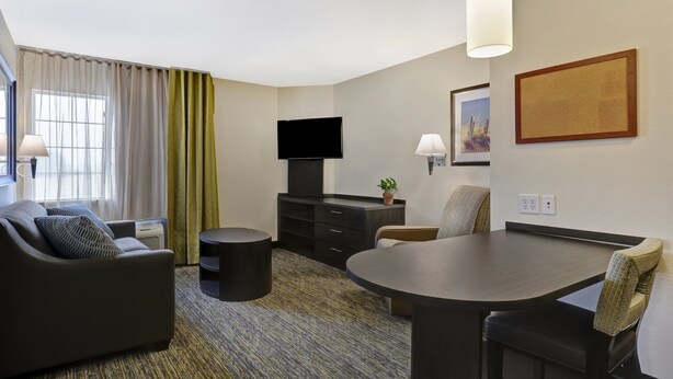 A modern hotel room with a sofa, a dining table, a TV, and a small side table. - Candlewood Suites Indianapolis - South by IHG (Greenwood)