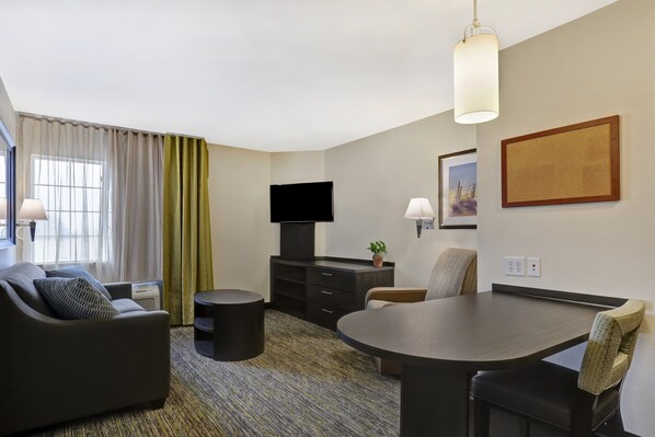 A modern hotel room with a sofa, a dining table, a TV, and a small side table. - Candlewood Suites Indianapolis - South by IHG (Greenwood)