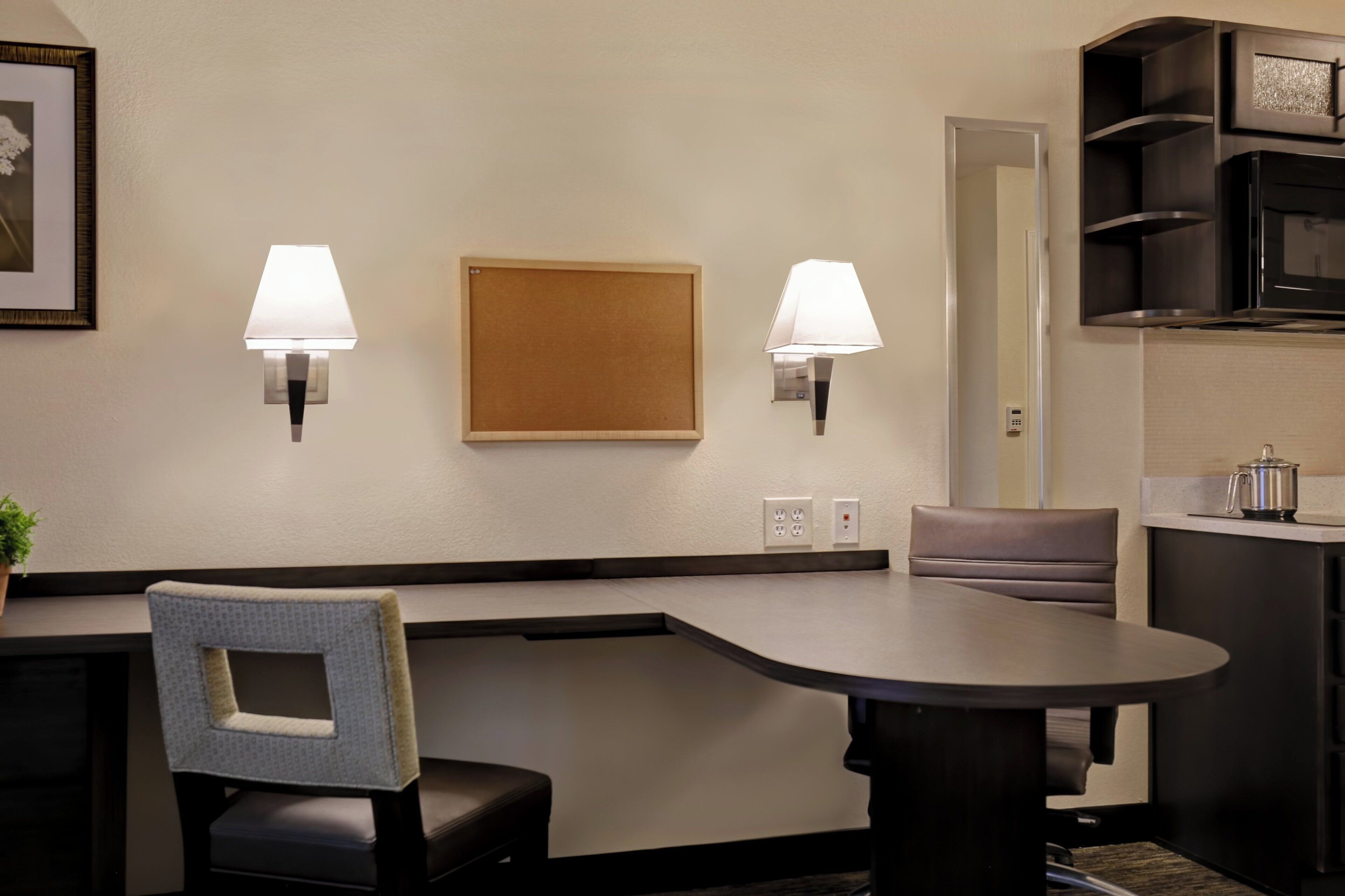 A hotel room with a dining table, two wall-mounted lamps, a framed picture, a sofa, and a kitchenette area.