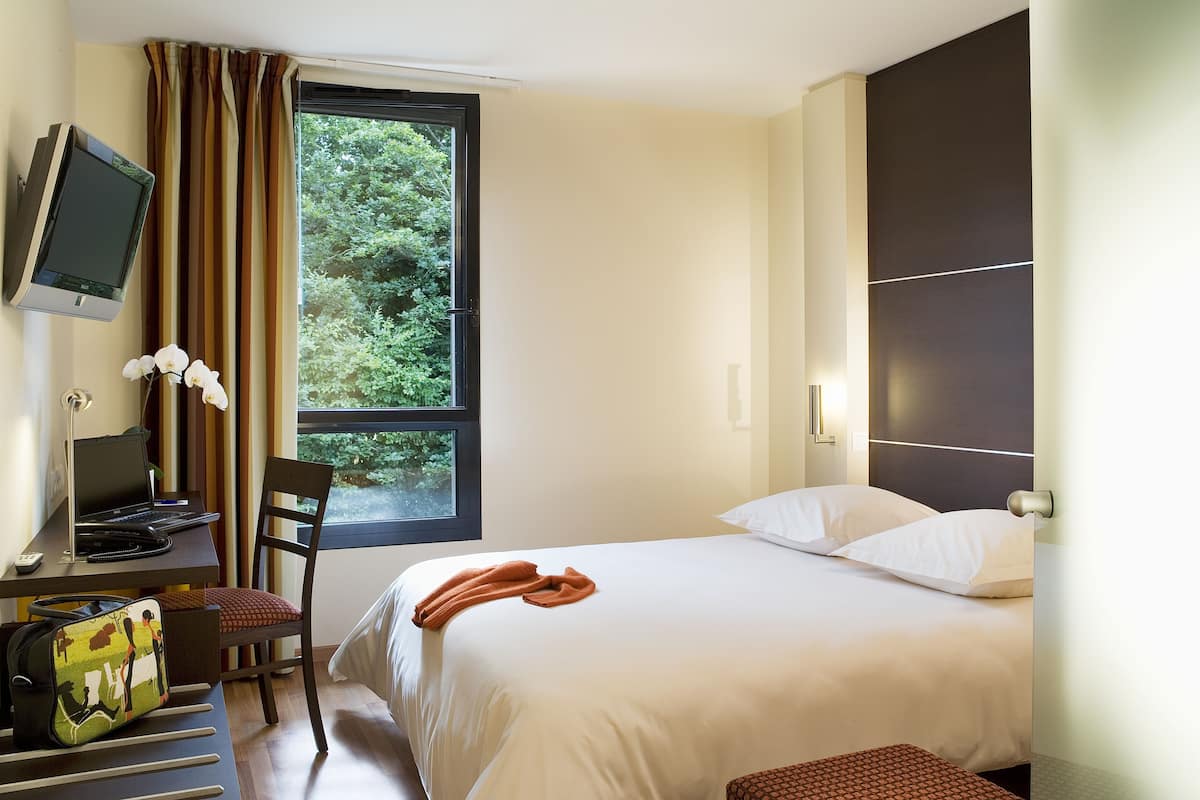 A hotel room with a bed, a desk with a laptop, a chair, a TV mounted on the wall, and a window with curtains.